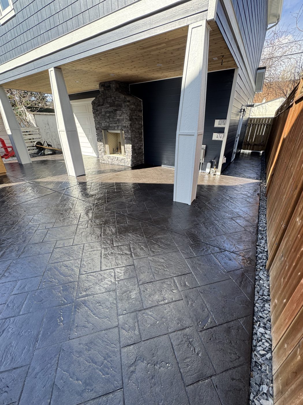 Charcoal slate-pattern stamped concrete patio with a stone outdoor fireplace under a covered lower deck in Coventry Hills, Calgary by Patriarch Construction